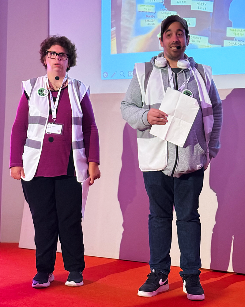 Två personer från Vår röst står på scen under LSS i fokus 2026, iförda reflexvästar och headsetmikrofoner. Bakom dem visas en presentation med en karta över Skåne med orter och siffror. Personerna håller i pappersark medan de berättar om sitt projekt.